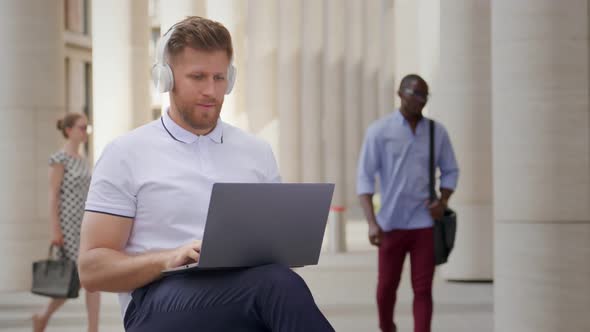 Cheerful Caucasian Man in Headphones Using Laptop Sitting on Staircase Outdoors