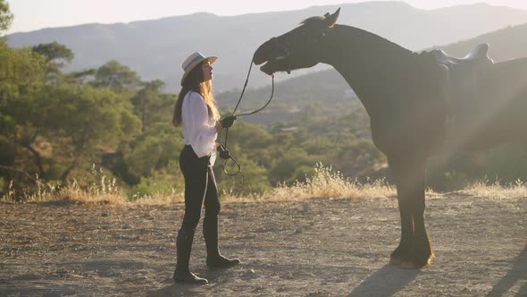 Side View of Gorgeous Brown Purebred Horse and Slim Confident Equestrian in Sunrays on Mountains