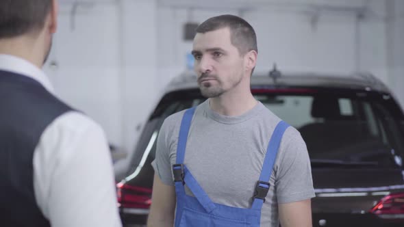 Portrait of Young Caucasian Auto Mechanic Talking with Customer in Repair Shop. Handsome Man 