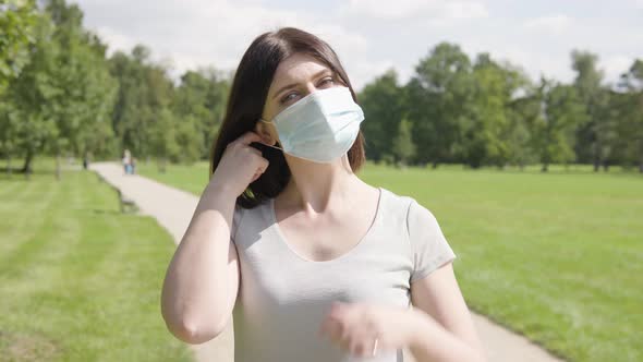 A Young Caucasian Woman Takes Off a Face Mask and Looks at the Camera on a Pathway in a Park