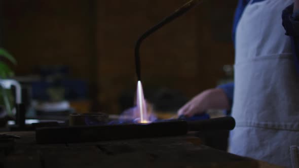 Midsection of caucasian female jeweller in workshop wearing apron, using gas burner