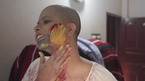 Happy and hopeful cancer patient bald girl applies colored powder on cheeks at home on Holi, Indian