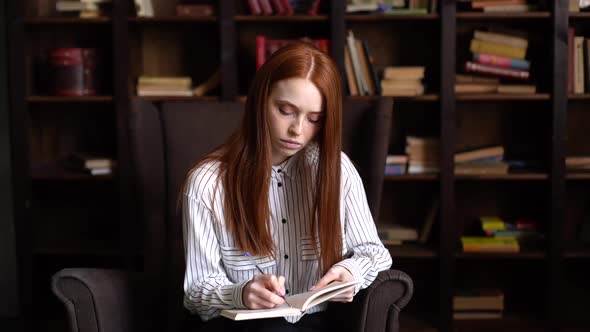 Focused Young Businesswoman with Red Hair Making Notes in Notebook at Home Office.