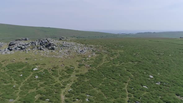 Wide aerial shot tracking forward over a tor surround by luscious green moorland. Sunny, Dartmoor, E