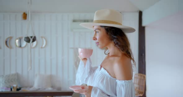 Young Charming Woman at Cozy Cafe Drinks Coffee From Pink Cup and Smiles with the Joy