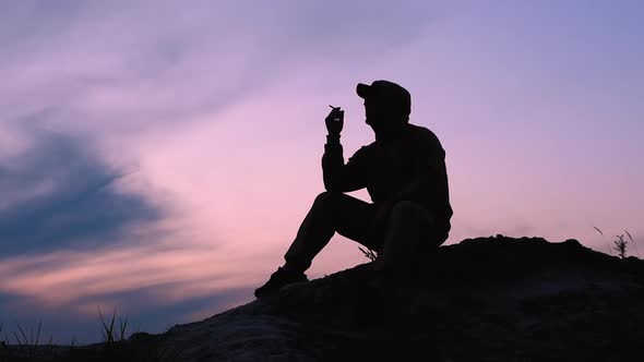 Silhouette Of A Smoking Man On Top Of A Mountain At Sunset. Breathing Out Cigarette Smoke