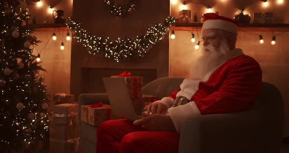 Santa Claus Is Working on a Laptop Sitting at Home on the Sofa in the Background of Christmas. Santa