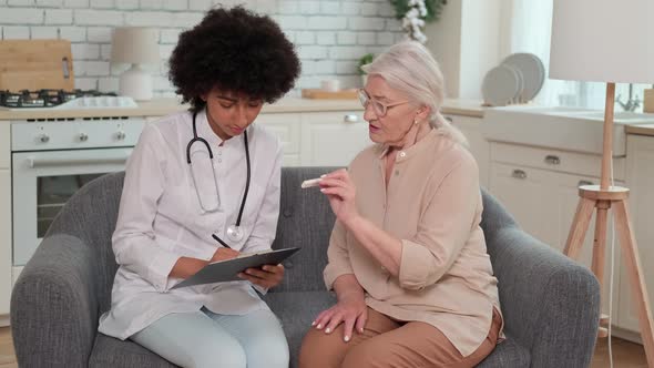 Afro American Woman Doctor Looking at Thermometer Reading at Senor Woman Sitting on Sofa at Home