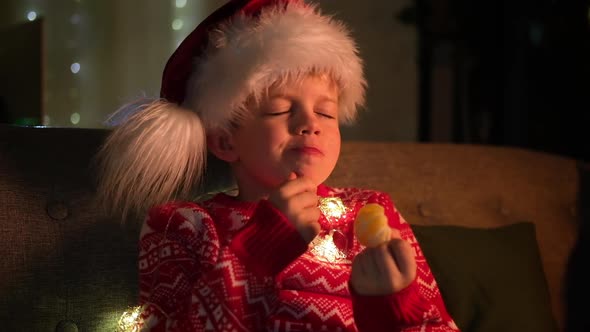 Portrait Dreaming Boy in Santa Hat and Christmas Costume Sitting at Home Eating Orange Tangerine