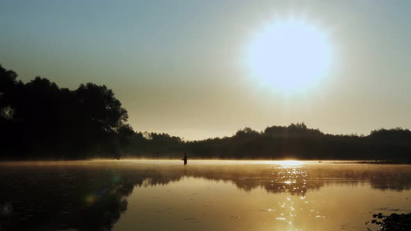 Silhouette of a fisherman on the river on a foggy summer morning. Dawn.