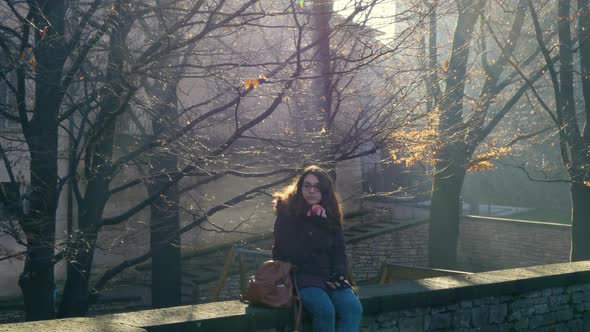 Young woman sitting on an old wall eats an apple while looking at the beautiful park. The rays of li