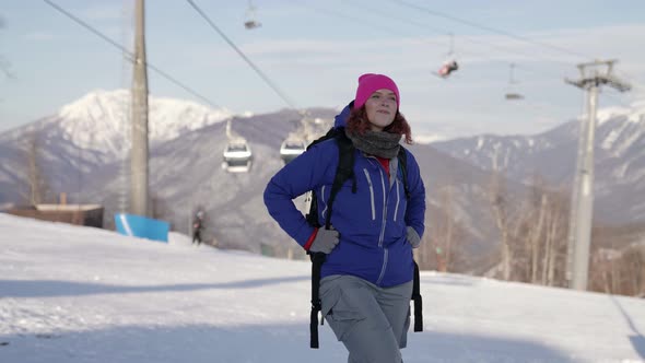 A Woman in a Pink Hat and Blue Jacket Stands Against the Backdrop of the Ski Lifts Smiling She Walks