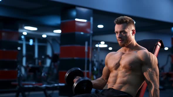 Athletic handsome man trains with dumbbells on fitness chair. Modern gym background. Slow motion.