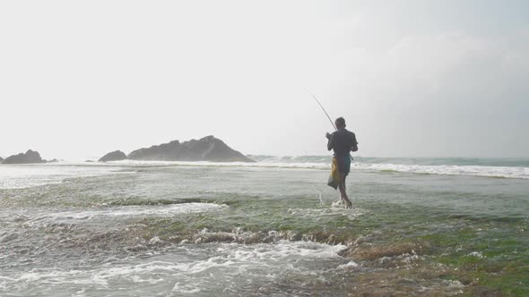 Aged Sinhalese Fisherman Walks in Surf Along Green Coastline