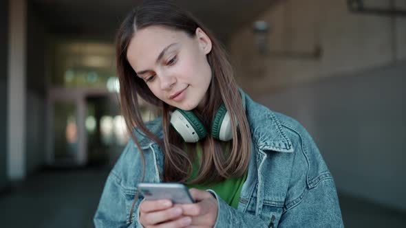 Concentrated blonde woman in headphones wearing jeans jacket typing by phone