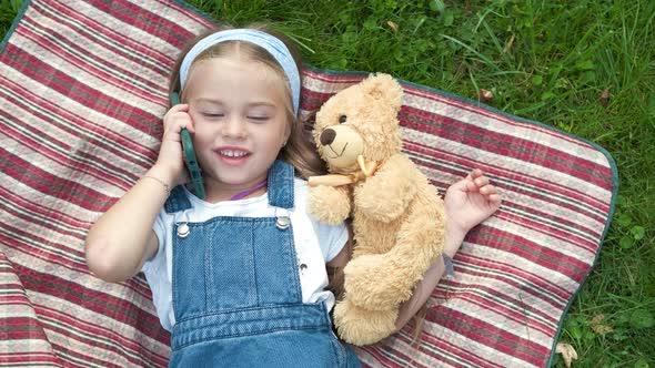 Little happy child girl laying on green lawn with her teddy bear talking on mobile phone.