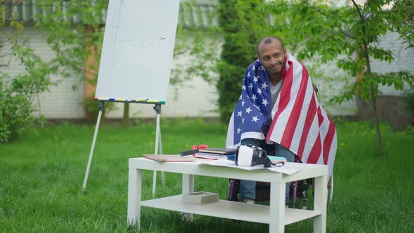 Confident Smiling Disabled Man in Wheelchair Covered with USA Flag Looking Around in Summer Garden