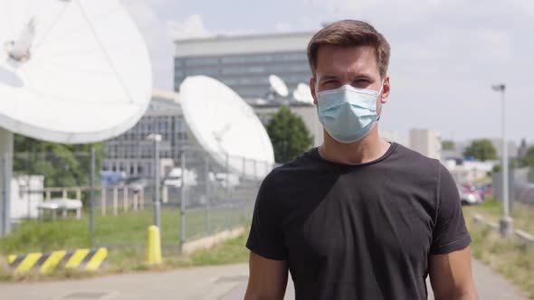 A Young Handsome Man in a Face Mask Looks at the Camera in an Urban Area - Satellite Dishes