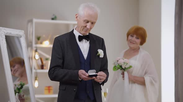 Proud Senior Caucasian Groom Posing with Wedding Rings As Happy Bride Standing at Background with