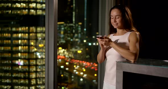 an Brunette with Long Hair Writes a Message in a Phone Near a Large Window. She Smiles and Thinks