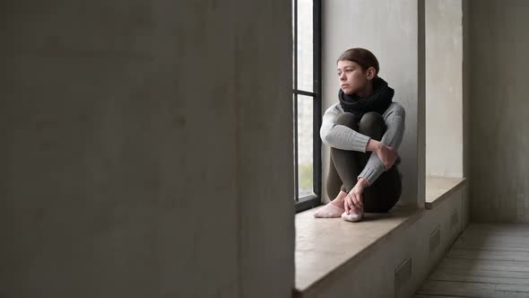 A sickly-looking young woman is sad on the windowsill of a city apartment