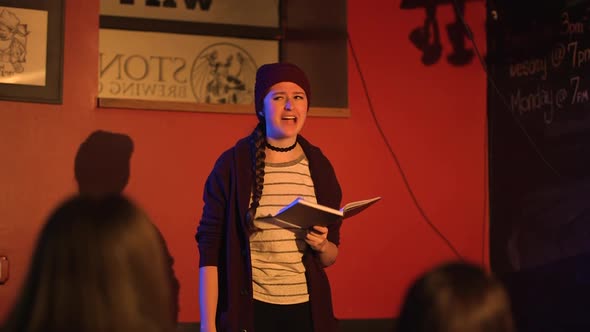 Young women reads slam poetry book on stage wearing red jacket and beanie