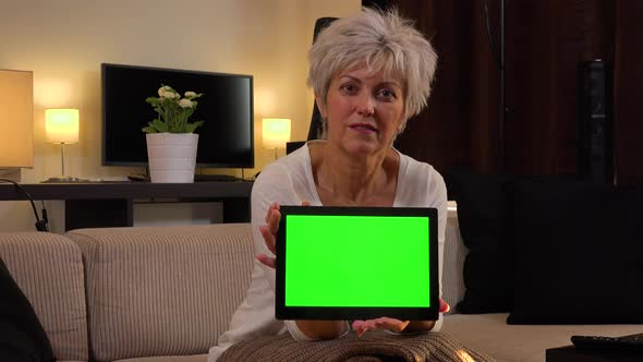 A Friendly Middle-aged Woman Sits and Shows a Tablet with a Green Screen To the Camera