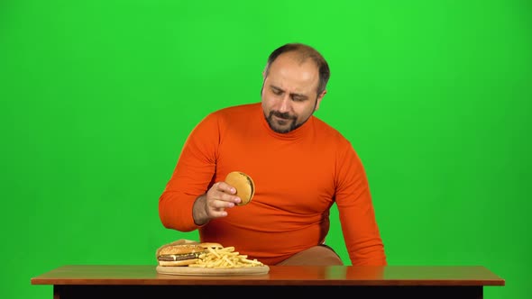 Caucasian Man at the Table Chooses Between a Plate of Fast Food and Healthy Food, Stops at Unhealthy