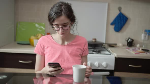 Nice Brunette Girl Sitting in the Kitchen Using Her Mobile Phone While Having Breakfast