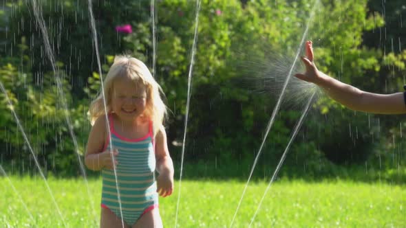 Two Happy Little Girls in Swimsuits are Playing with a Water Sprinkler on a Lawn