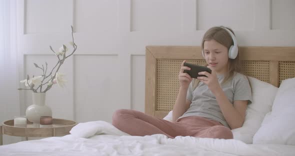 Child Girl Is Playing Game in Smartphone After School Lessons, Sitting on Bed at Home Alone