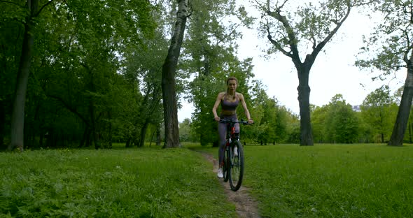 A Beautiful Slender Girl in a T-shirt and Leggings Rides a Bicycle Along the Path Among the Trees