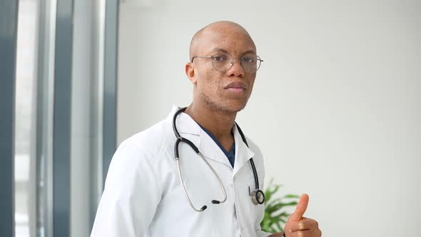 A Young African American Male Doctor Stands in a Medical Suit at the Clinic and Looks at the Camera