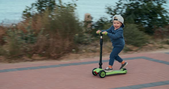 Little Boy in a Cap Quickly Rides a Green Scooter After His Mother Along the Sea