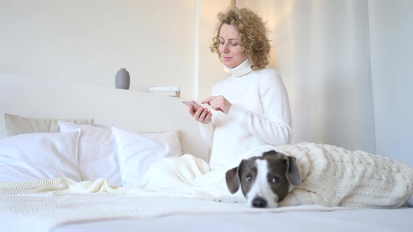 Young Woman Chatting On Mobile Phone On Bed With Dog In Cozy Sweater.