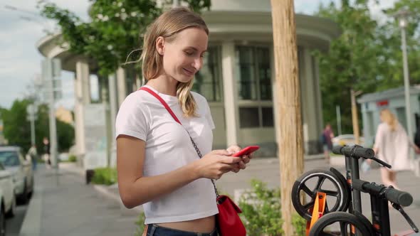 Woman Using Smartphone To Pay for Rental Electric Scooter While Standing on Parking Lot on Modern