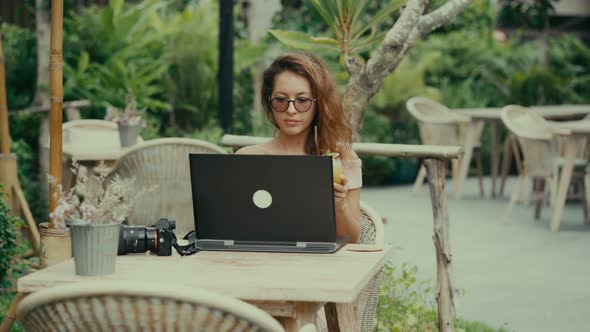 A Young Woman Photographer Drinks a Cocktail While Working with Her Laptop in an Outdoor Cafe