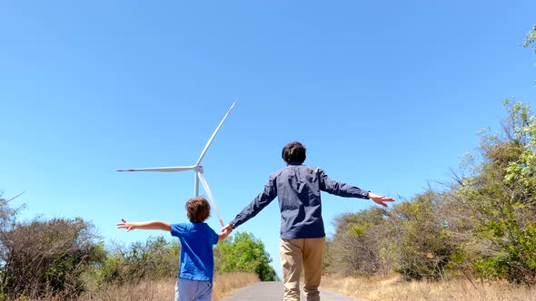 Slowmotion Shot of a Father and Son Running on a Road Leading to the Wind Electric Generator