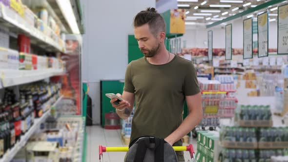 Young Male is Using Mobile Phone Choosing Beer at Supermarket