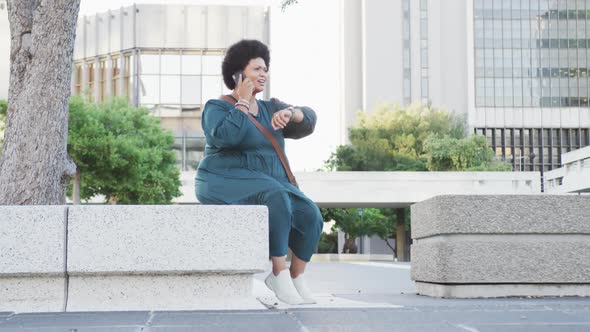 Happy plus size biracial woman talking on smartphone and checking smartwatch