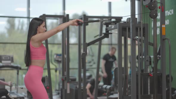 A Brunette Woman in a Pink Suit Pulls a Rope in a Crossover Trainer to Her Chest