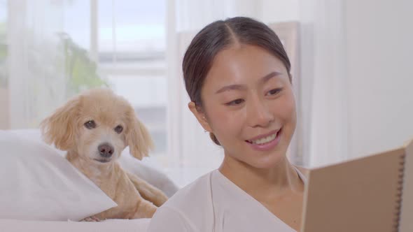 Beautiful Asian young woman reading book with dog in cozy bedroom relax and comfortable at home