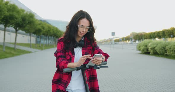 Brunette in Glasses and with Headphones Stopped During Walking and Using Her Smartphone