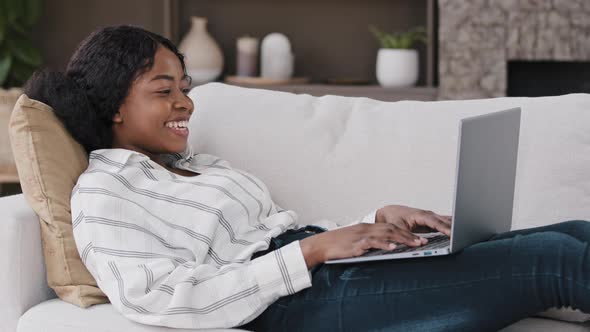 African Woman Lying on Sofa at Home Look at Laptop Screen Greeting Hello Make Video Call Talking