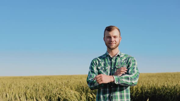 A Young Farmer Agronomist with a Beard Stands in a Wheat Field Under a Clear Blue Sky