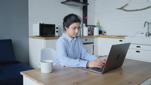 Indian Woman Working at a Computer Online Sitting at a Table on a chair in the Kitchen