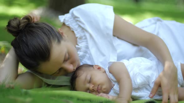 Mother Kisses Her Baby Sleeping on a Green Grass Outdoors. Happy Smiling Young Mother and Child in