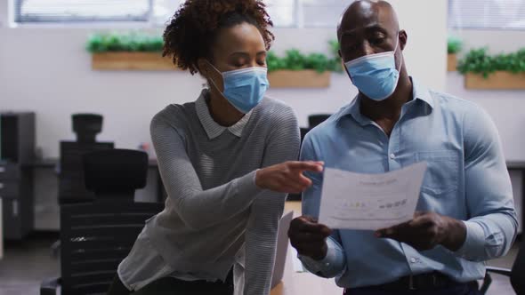 Diverse male and female business colleagues in face masks talking, man holding document in office