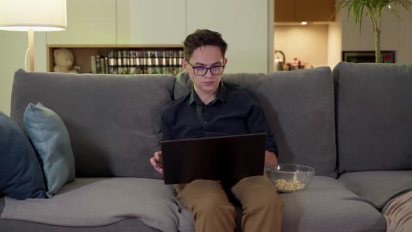 A Young Guy with Glasses Sits on a Sofa with a Laptop