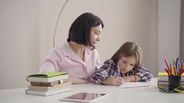 Young Caucasian Mother Dictating To Daughter As Pretty Brunette Girl Writing Down. Schoolchild Doing